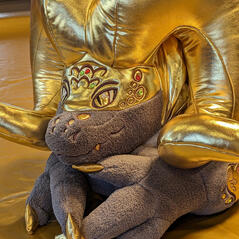 A photograph of a fantastical plush lizard with golden ram's horns laying on a gold colored cloth.