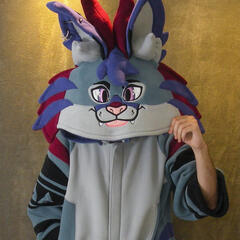 A photograph of the artist wearing a kigurumi styled after a steel gray lynx with red and purple tufts of fur.