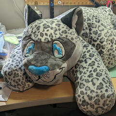 A photograph of a giant plush snow leopard laying on a cluttered sewing table.