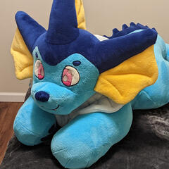 A photograph of a plush frilly axolotl/cat hybrid laying on a cushion.